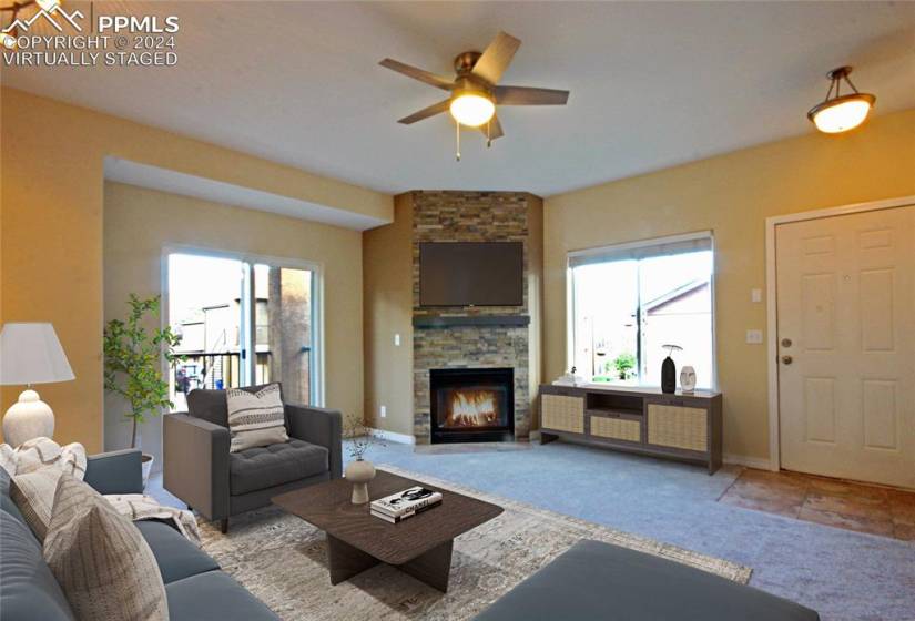 Living room with a fireplace, brick wall, ceiling fan, and light carpet, virtually staged