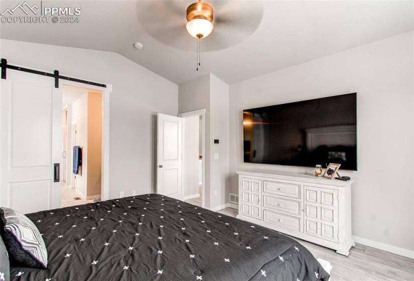 Bedroom with ensuite bathroom, a barn door, light hardwood / wood-style flooring, vaulted ceiling, and ceiling fan