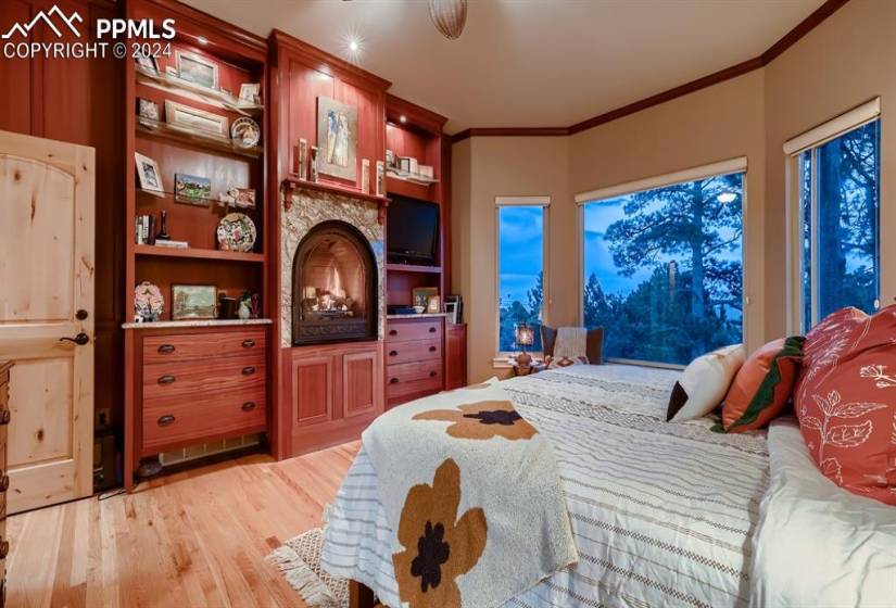 Primary bedroom featuring light hardwood floors, built ins, a stone fireplace and crown molding