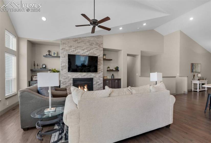 Living room featuring ceiling fan, built in shelves, hardwood flooring, high vaulted ceiling, and a stone fireplace