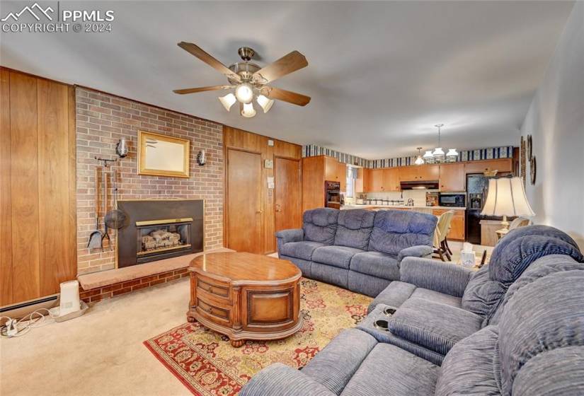 Carpeted living room featuring brick wall, ceiling fan with notable chandelier, a fireplace, and wood walls