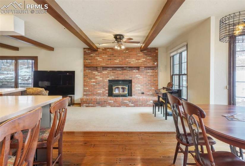Carpeted living room with a brick fireplace, ceiling fan, beamed ceiling, and a healthy amount of sunlight