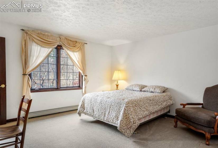 Bedroom with it's own bathroom, baseboard heating unit, light colored carpet, and a textured ceiling