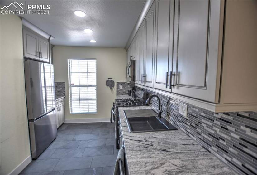 Kitchen with dark tile floors, sink, backsplash, appliances with stainless steel finishes, and light stone countertops