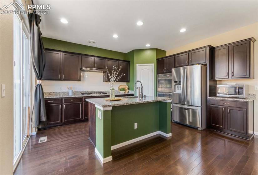 Kitchen with a kitchen island with sink, appliances with stainless steel finishes, dark hardwood / wood-style floors, and light stone counters