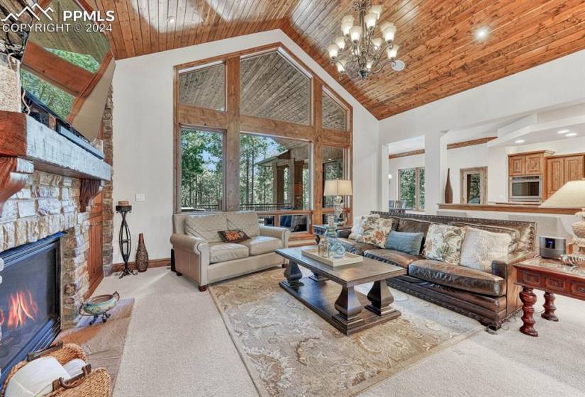 Carpeted living room featuring a chandelier, high vaulted ceiling, a stone fireplace, and a healthy amount of sunlight and pine trees