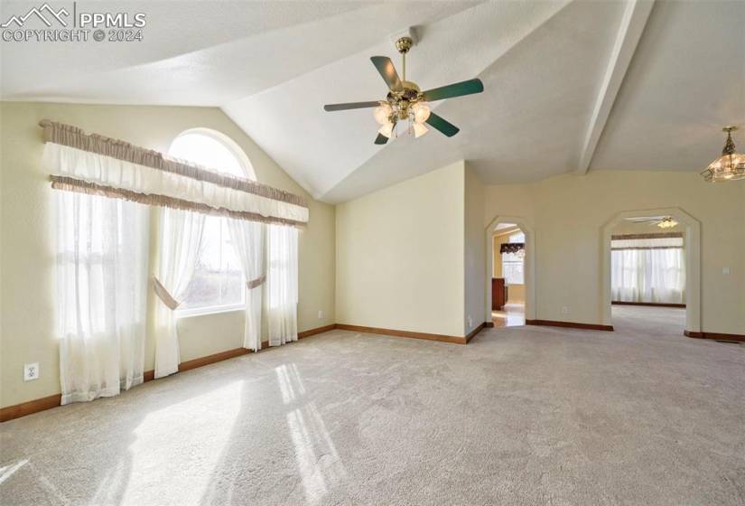 Carpeted room with vaulted ceiling with beams, ceiling fan with notable chandelier, and plenty of natural light