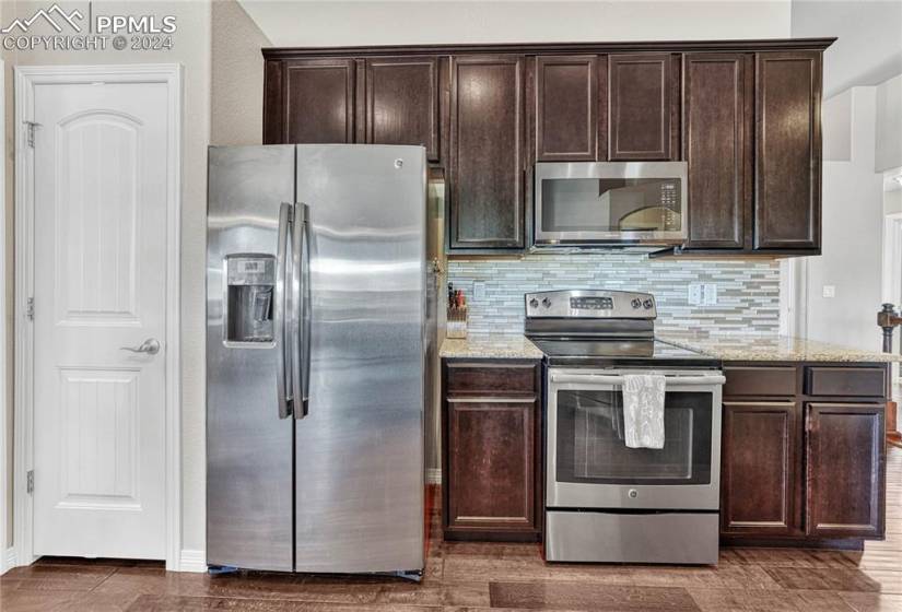 Kitchen featuring backsplash, appliances with stainless steel finishes, dark hardwood / wood-style floors, dark brown cabinetry, and light stone countertops
