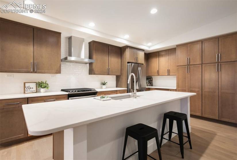 Kitchen featuring a kitchen breakfast bar, wall chimney range hood, a center island with sink, appliances with stainless steel finishes, and light hardwood / wood-style flooring