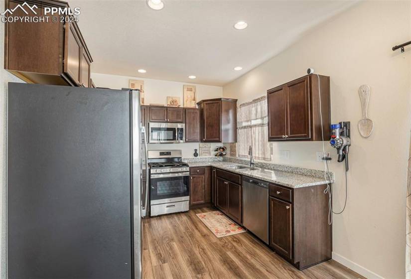 Kitchen with sink, appliances with stainless steel finishes, light wood-type flooring, light stone counters, and dark brown cabinets