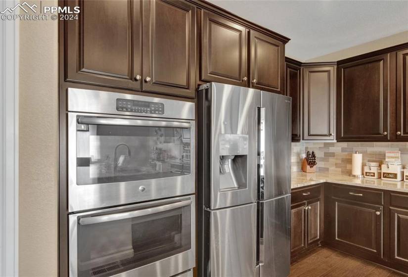 Kitchen featuring backsplash, stainless steel appliances, double ovens, granite counters, dark engineered hardwood floors, light stone counters, and dark brown cabinetry