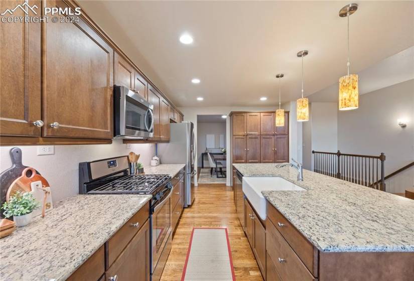 Kitchen with stainless steel appliances, a kitchen island with sink, hanging light fixtures, light wood-type flooring, and light stone countertops