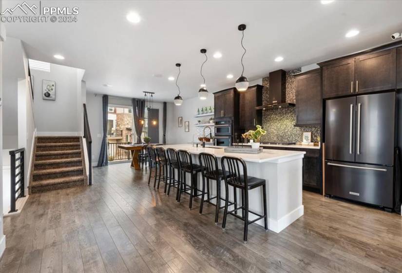 Kitchen with hardwood / wood-style flooring, stainless steel appliances, a kitchen island with sink, dark brown cabinetry, and wall chimney exhaust hood
