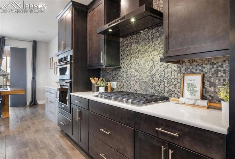 Kitchen featuring wall chimney range hood, tasteful backsplash, stainless steel appliances, light hardwood / wood-style flooring, and dark brown cabinetry