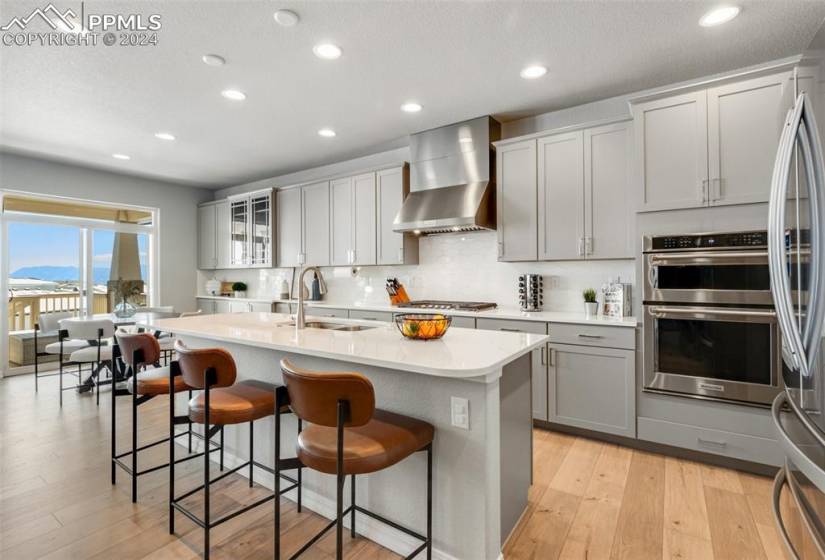 Kitchen with sink, a kitchen island with sink, appliances with stainless steel finishes, light wood-type flooring, and wall chimney range hood