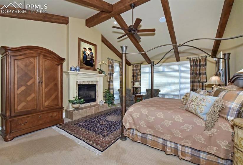 Carpeted bedroom with a tiled fireplace, vaulted ceiling with beams, and ceiling fan