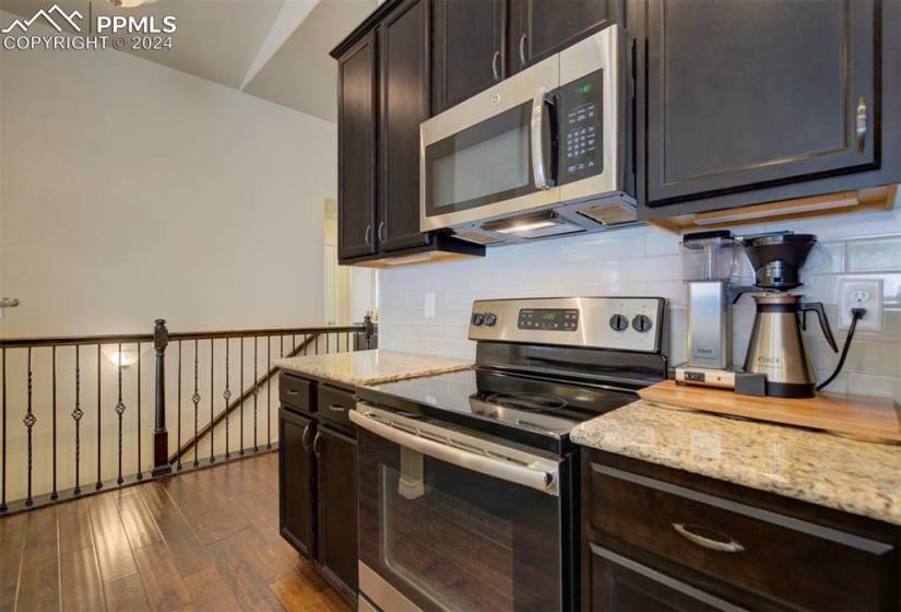 Kitchen with stainless steel appliances, tasteful backsplash, dark brown cabinets, dark wood-type flooring, and light stone counters