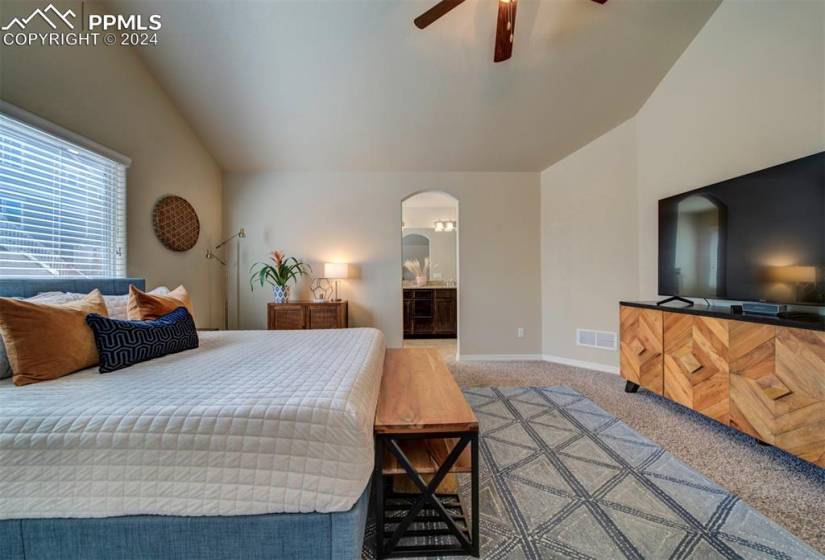 Carpeted bedroom featuring ceiling fan, ensuite bath, and vaulted ceiling