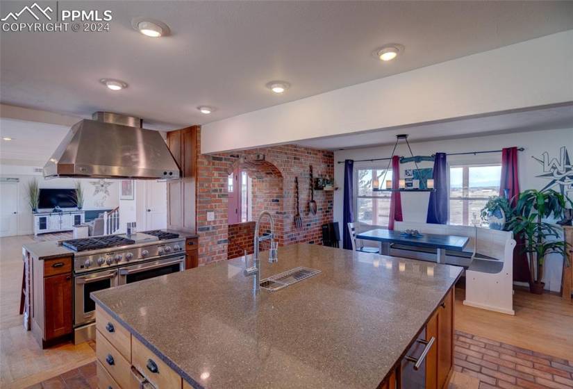 Kitchen featuring exhaust hood, double oven range, a center island with sink, brick wall, and dark stone counters