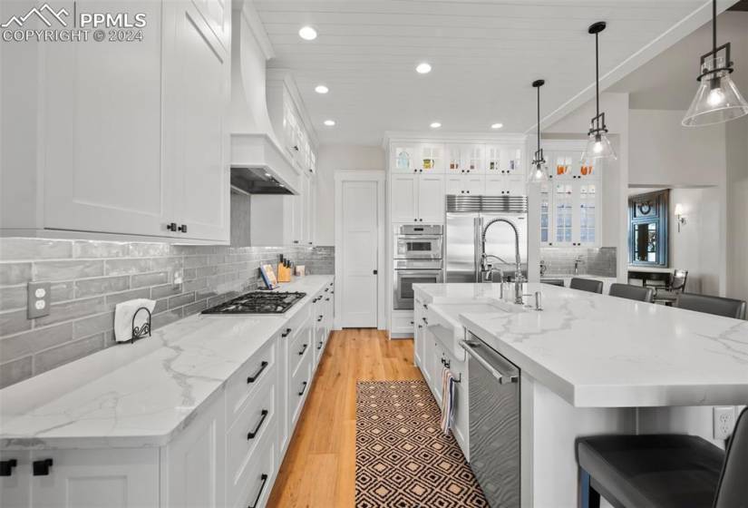 Kitchen with custom exhaust hood, hanging light fixtures, light wood-type flooring, appliances with stainless steel finishes, and a breakfast bar area