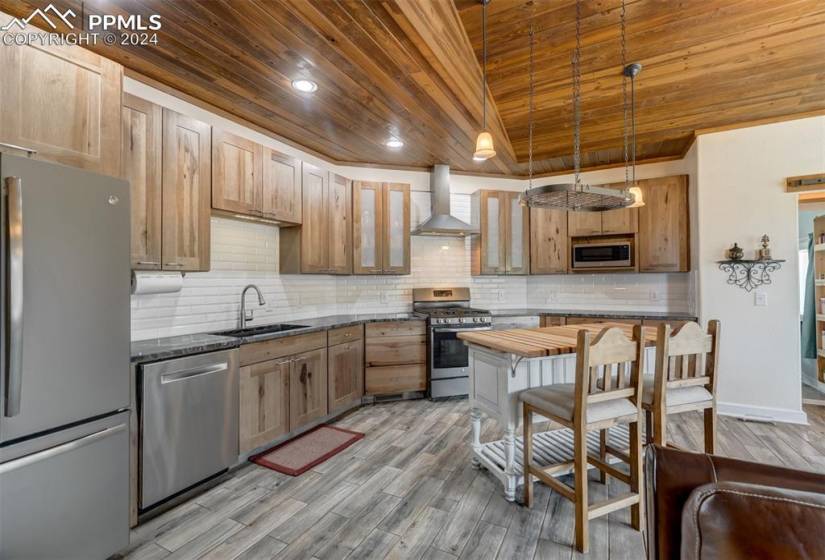 Kitchen featuring wall chimney range hood, light wood-type flooring, tasteful backsplash, stainless steel appliances, and wooden ceiling
