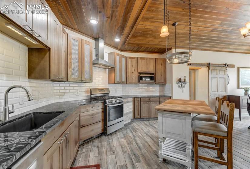 Kitchen featuring wall chimney range hood, backsplash, wood-type flooring, stainless steel appliances, and wooden ceiling