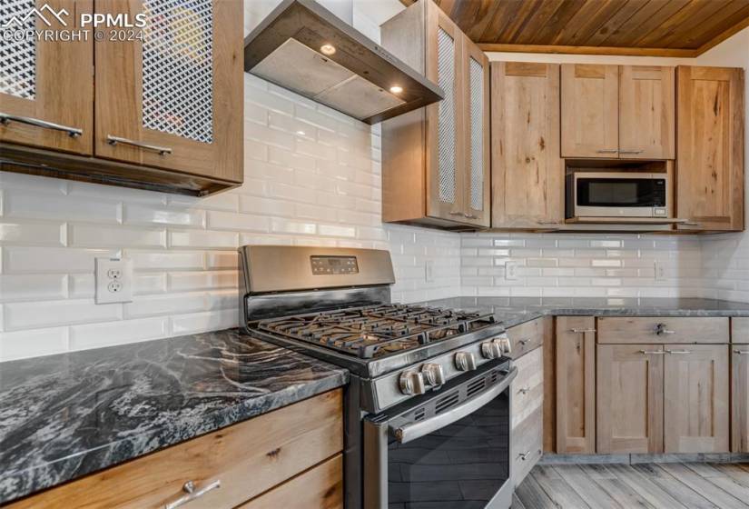 Kitchen featuring wall chimney range hood, stainless steel appliances, tasteful backsplash, dark stone countertops, and light hardwood / wood-style floors