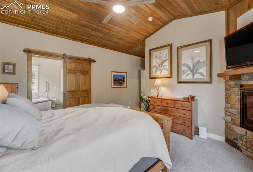 Carpeted bedroom featuring a barn door, a stone fireplace, ceiling fan, wood ceiling, and connected bathroom
