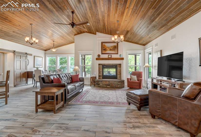 Living room with a stone fireplace, high vaulted ceiling, ceiling fan with notable chandelier, hardwood / wood-style floors, and wooden ceiling