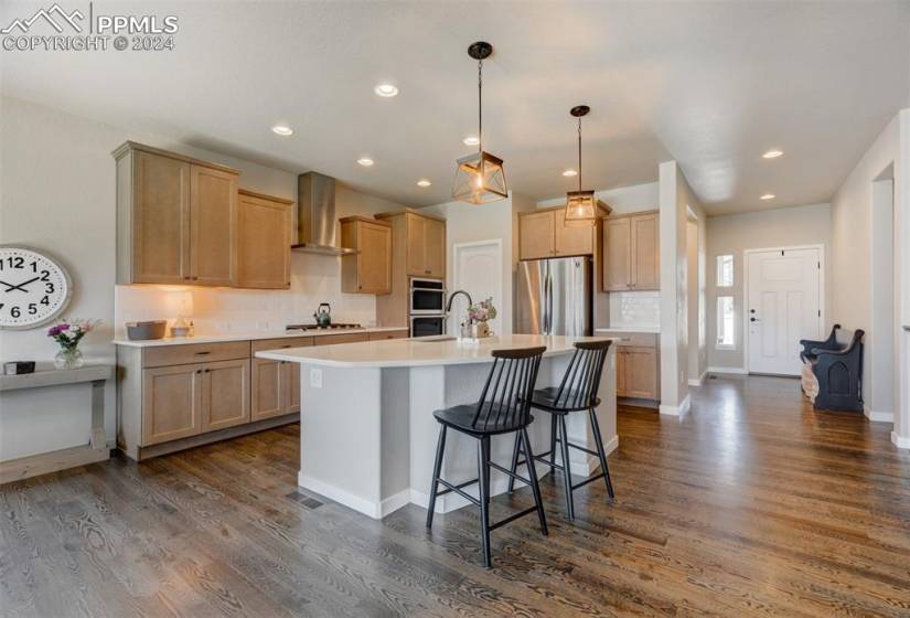 Kitchen featuring wall chimney exhaust hood, dark hardwood / wood-style flooring, backsplash, appliances with stainless steel finishes, and an island with sink
