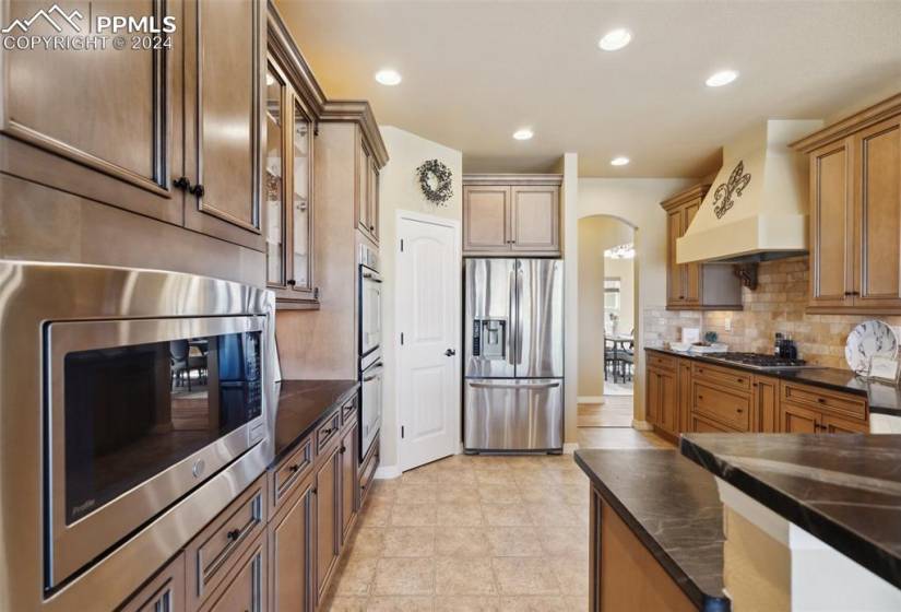 Kitchen featuring stainless steel appliances, premium range hood, tasteful tile backsplash, dark stone counters, and tile floors
