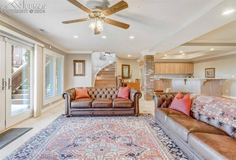 Tiled living room with crown molding, a tray ceiling, brick wall, ornate columns, and ceiling fan